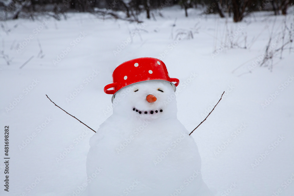 Funny snowman in a red soup pot with white dots instead of a hat, a cute snowman stands in a winter village, snow-covered trees