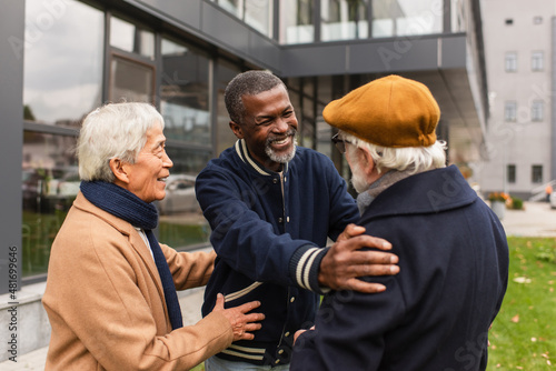 Fototapeta Naklejka Na Ścianę i Meble -  Happy african american pensioner hugging senior friend near asian man on urban street.