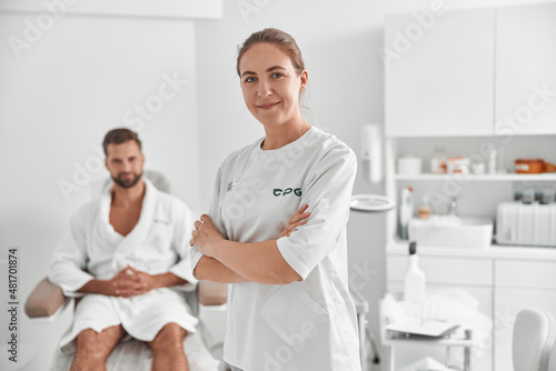Portrait of positive cosmetologist in white uniform against blurred view of man patient in beauty salon