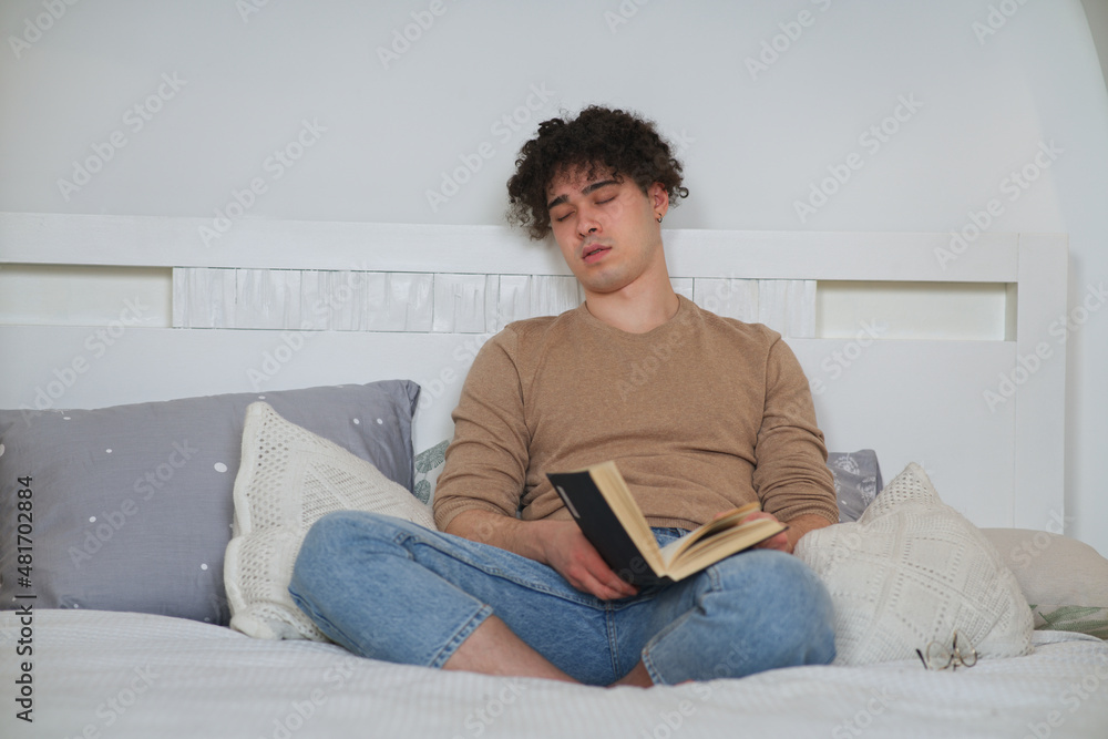 Portrait of handsome tired exhausted sleepy guy, young man is sleeping sitting on cozy bed in bedroom with open book in hands. Reader fall asleep, daydream at home. Student preparing to exam
