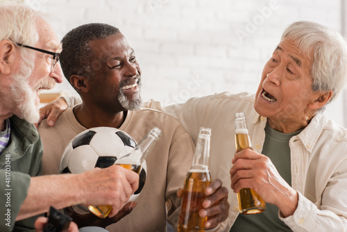 Excited interracial senior friends holding beer bottles and football at home.