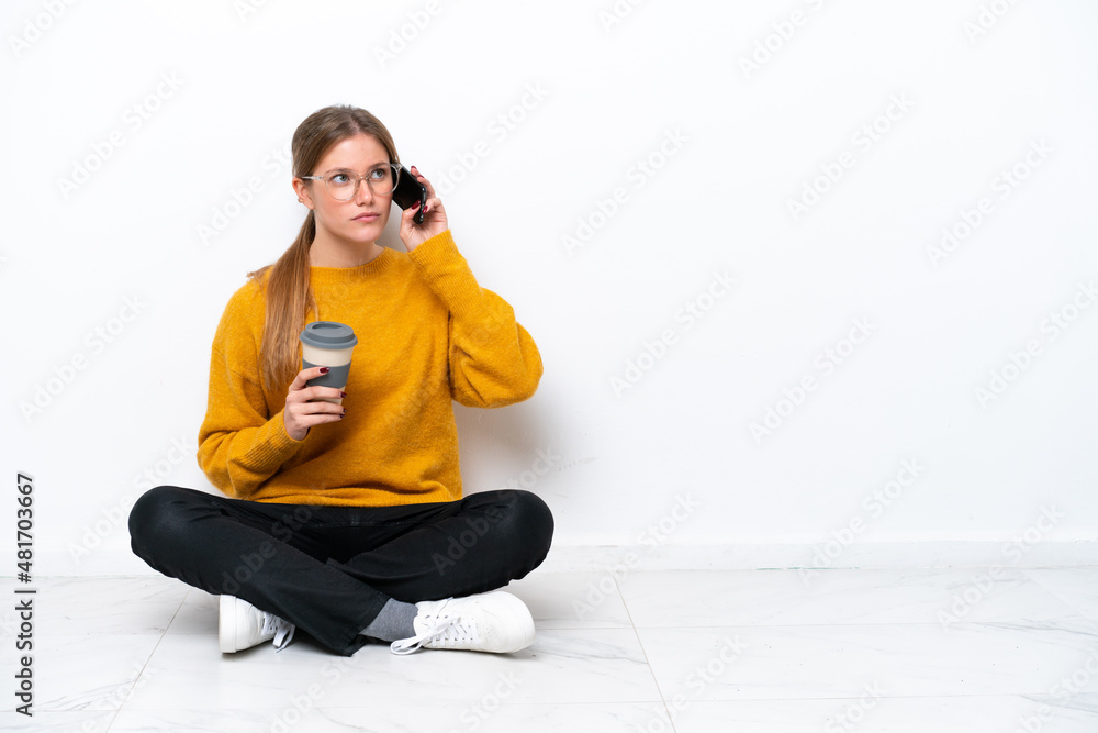 Young caucasian woman sitting on the floor isolated on white background holding coffee to take away and a mobile Young caucasian woman sitting on the floor isolated on white background holding coffee to take away and a mobile