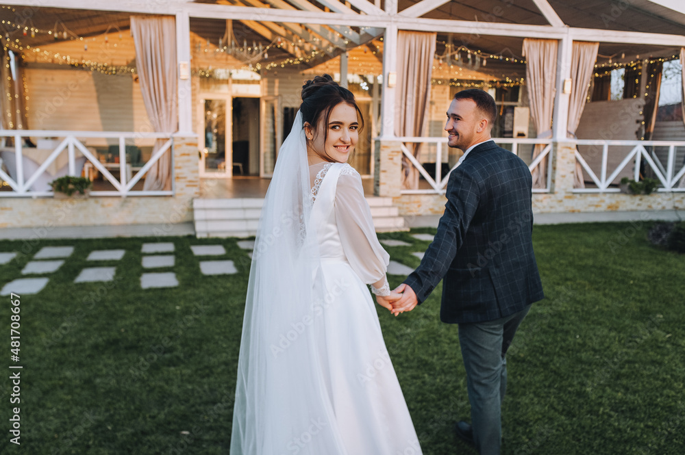 Fototapeta premium A young groom and a beautiful, smiling brunette bride in a white dress are walking along the green grass, in nature, against the backdrop of a cottage, at home. Wedding portrait, photography.