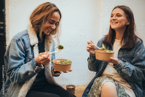 Two young women female friends sitting outdoors eating takeaway food, laughing and having fun. Food delivery and takeout.