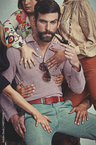 I'm a ladies man, for sure. Studio shot of an attractive man in retro 70s wear being touched by women while smoking his pipe. photo