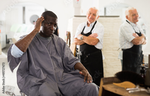 African-American man sitting in hairdressing chair, gazing at his haircut performed by experienced aged barber