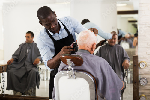 Skillful African man barber making hairstyle for senior male client in hair studio, using electric trimmer
