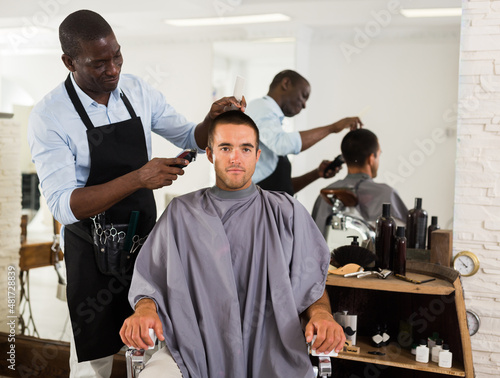 Male client getting trendy haircut at barber shop from African-American hairstylist using machine