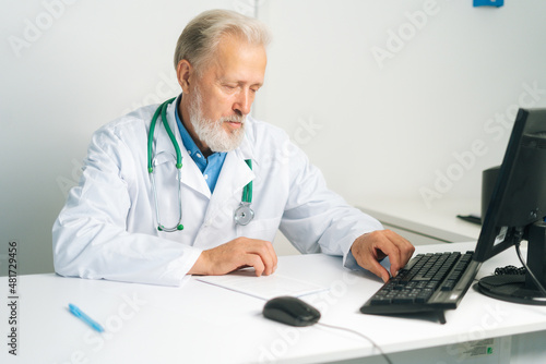 Portrait of focused mature adult male doctor wearing white coat with stethoscope working on desktop computer sitting at desk in medical office. Confident senior physician at workplace in hospital.