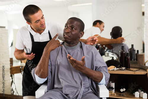 Portrait of dissatisfied African male client in hair salon with perplexed hairdresser..
