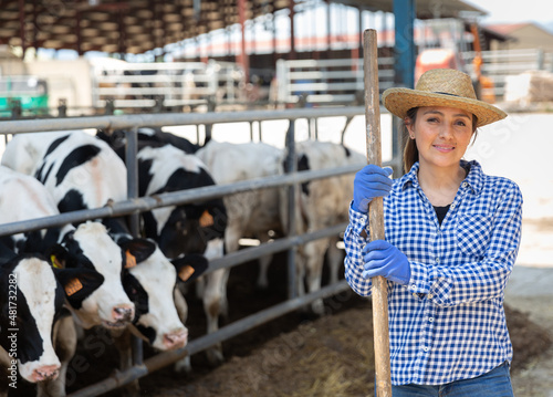 Positive young hispanic woman owner of dairy farm standing in stall on background with herd of cows