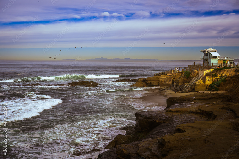 2022-01-19 THE LIFEGUARD STATION ALONG THE ROCKY SHORE IN LA JOLLA WITH WAVES CRASHING AND BIRDS IN A CLOUDY SKY
