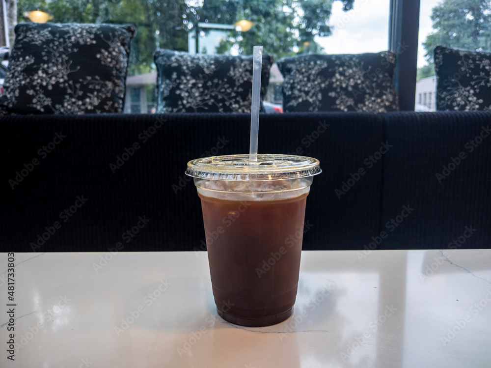 Iced, cold brew coffee in a plastic cup with straw, sitting on a white table in a cozy cafe with black padded bench seating Iced, cold brew coffee in a plastic cup with straw, sitting on a white table in a cozy cafe with black padded bench seating
