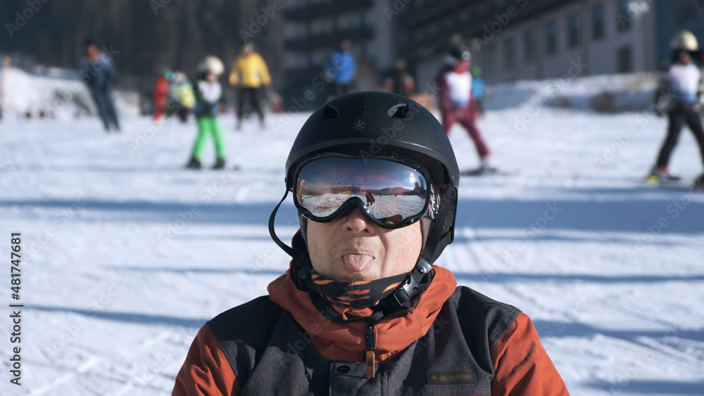 Close-up, funny man snowboarder portrait, on snow-covered mountain slope, backdrop of people skiing on sunny winter day. Happy man looking, smiling at camera in ski mask, helmet. Play active sports Close-up, funny man snowboarder portrait, on snow-covered mountain slope, backdrop of people skiing on sunny winter day. Happy man looking, smiling at camera in ski mask, helmet. Play active sports