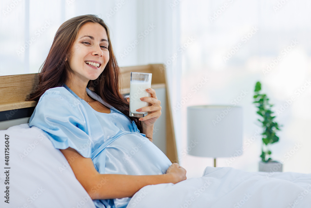 Cheeful pregnant woman smile as enjoy drinking glass of milk beverage in morning while resting on bed with lovely bear doll Cheeful pregnant woman smile as enjoy drinking glass of milk beverage in morning while resting on bed with lovely bear doll