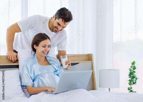 Lovely pregnant wife tenderly touch beloved belly of unborn baby while lounging on bed with smile and happy as enjoy watching entertainment media on computer