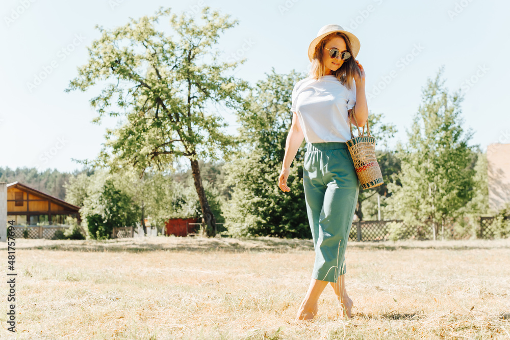 Charming caucasian cute young woman model in straw hat and glasses posing outdoors on summer sunny day. Slim smiling pretty girl standing barefoot on tiptoe in meadow, walking in nature