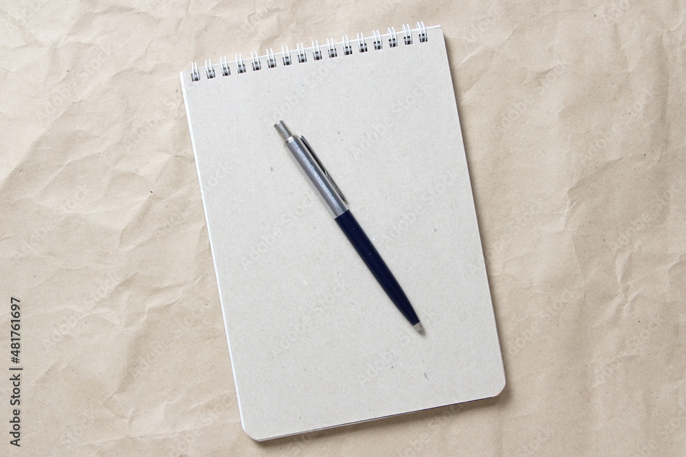Gray notepad with white coiled spring and pen on a background of beige crumpled craft paper Gray notepad with white coiled spring and pen on a background of beige crumpled craft paper
