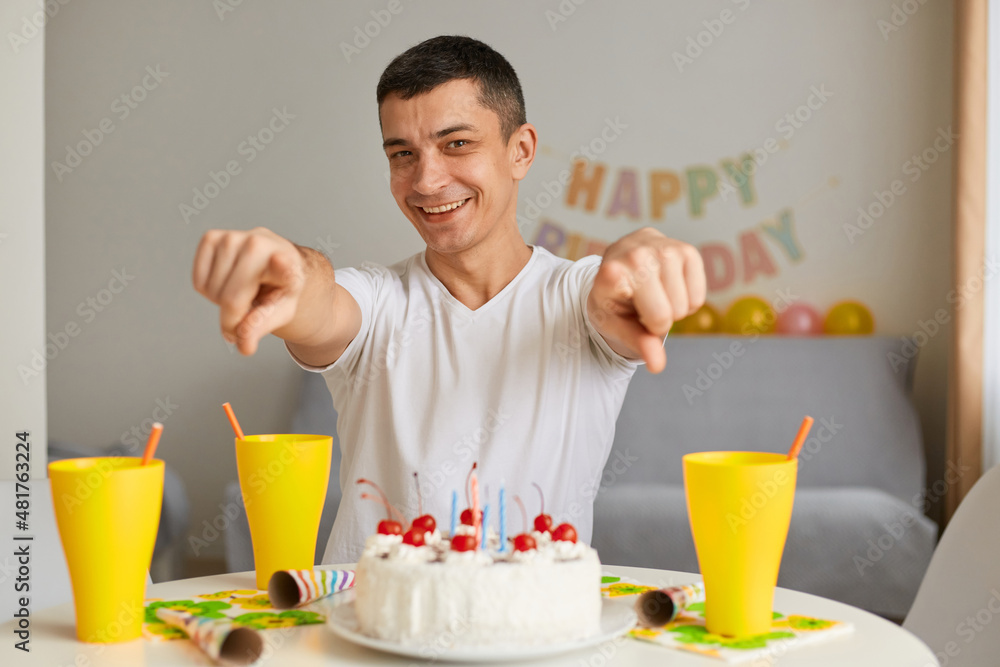 Portrait of smiling optimistic young man wearing white casual style T-shirt sitting at table with cake, pointing finger to camera, inviting you to his birthday party. Portrait of smiling optimistic young man wearing white casual style T-shirt sitting at table with cake, pointing finger to camera, inviting you to his birthday party.