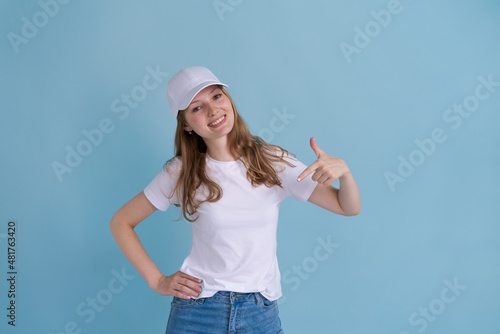 Young caucasian happy girl in delivery uniform and white cap looks confident with a smile on her face, pointing her fingers proudly and happily on a blue background © Екатерина Переславце