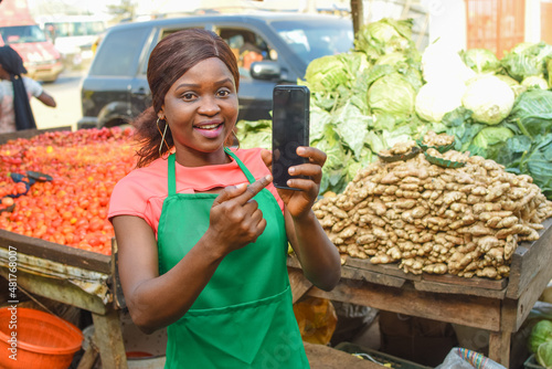 Happy African business woman or female trader wearing a green apron and pointing to a smart phone while standing at her vegetable stall in a market place