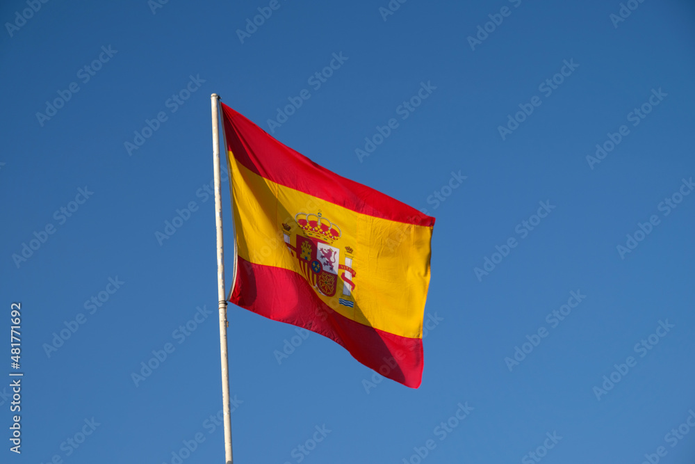 Spanish national flag waving on blue sky background. The flag on a flagpole flutters in the wind. Red with yellow Spain Flag blowing in the wind against on blue sky Spanish national flag waving on blue sky background. The flag on a flagpole flutters in the wind. Red with yellow Spain Flag blowing in the wind against on blue sky