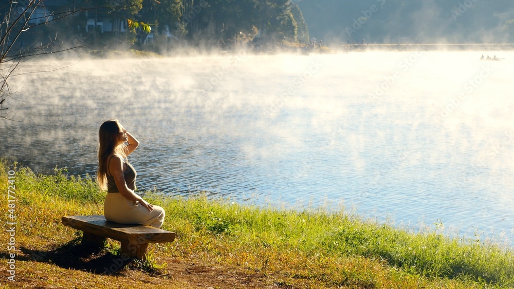 Fototapeta premium Travel woman relax on nature near misty lake. Female traveler sitting on bench, enjoy fog morning on river. Calmness, relaxation, meditation, freedom, beauty of nature concept