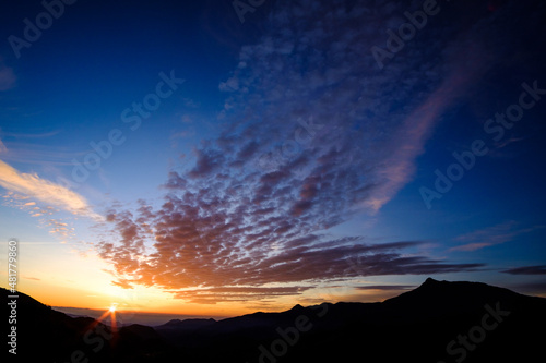 Orange cirrus clouds on a blue sunrise landscape form the top of a mountain