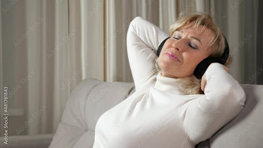 Blonde long-haired woman of middle age with bangs wearing light turtleneck listens to music via wireless headphones relaxing on sofa, close-up. Blonde long-haired woman of middle age with bangs wearing light turtleneck listens to music via wireless headphones relaxing on sofa, close-up.