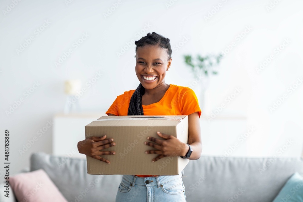 Overjoyed black lady holding cardboard parcel, receiving desired delivery, getting her online order at home - obrazy, fototapety, plakaty 