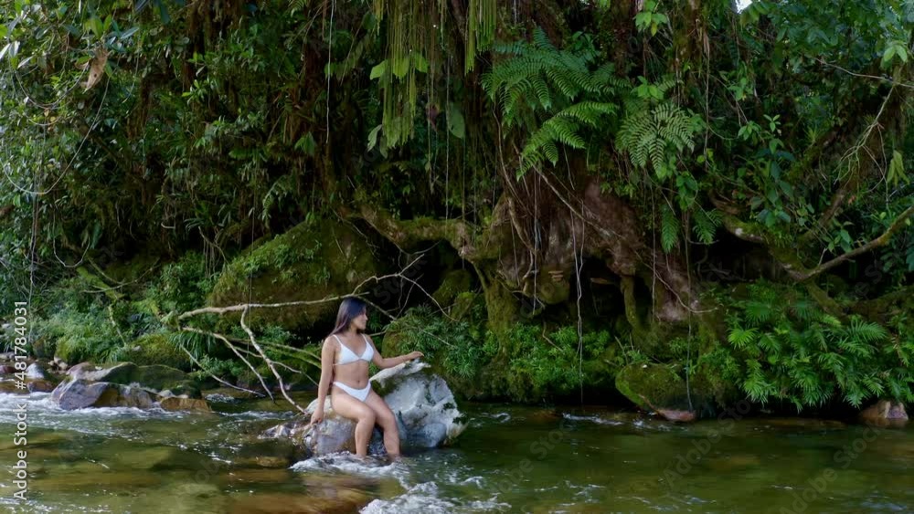 A beautiful woman with long black hair in a white bikini of hispanic ethnicity, is sitting and relaxing on a large rock in a river in nature A beautiful woman with long black hair in a white bikini of hispanic ethnicity, is sitting and relaxing on a large rock in a river in nature