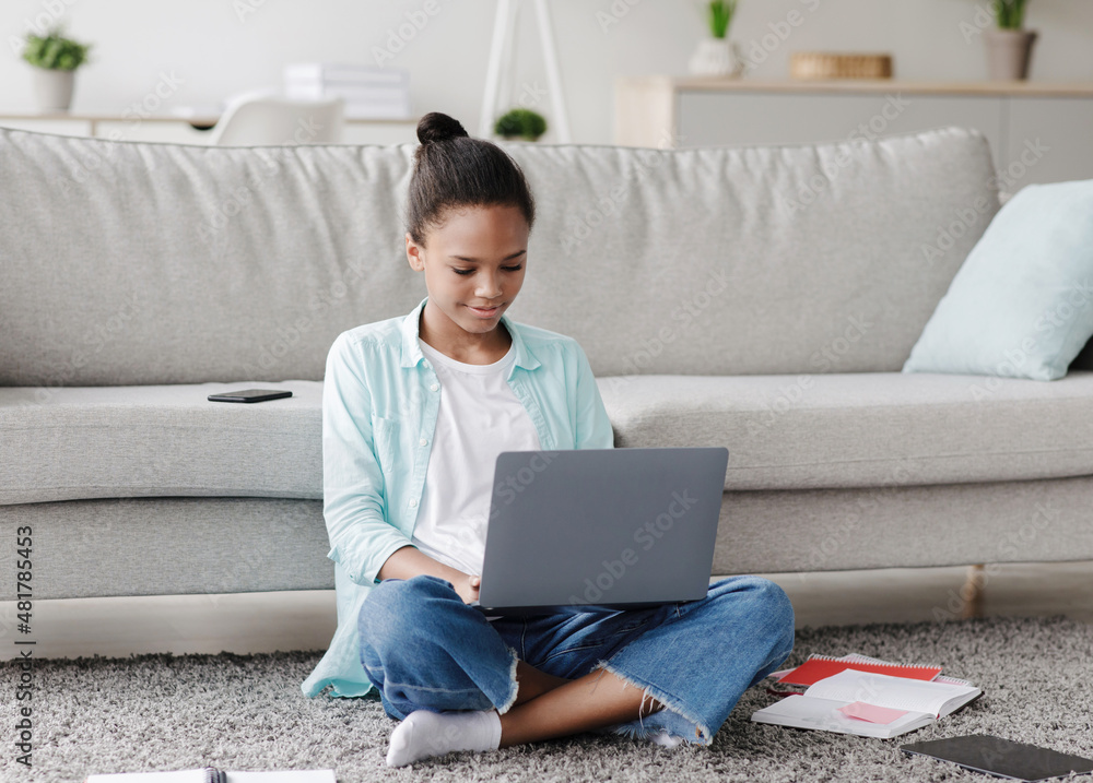 Naklejka premium Busy concentrated teen afro american girl studying remotely, sitting on floor and look at laptop in living room