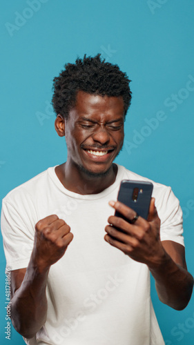 African american man feeling excited and using smartphone in studio. Black person looking at phone display and feeling cheerful, celebrating online success while browsing internet.