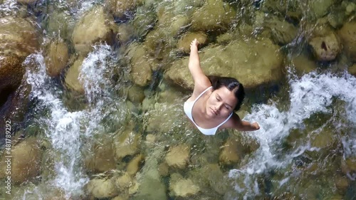 A woman is relaxing with her eyes closed in a river with crystal clear water, the woman has a white bikini and long black hair of hispanic ethnicity