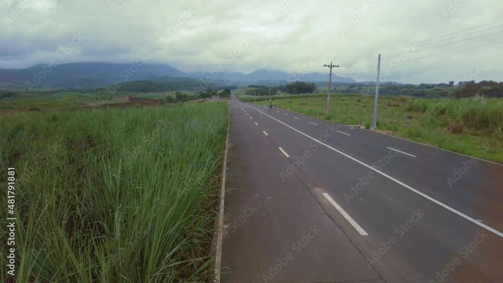 A jib shot of an empty road, surrounded by tall grass. This is also a sugarcane field with mountains beyond. Two bikers drives through from right to left. A jib shot of an empty road, surrounded by tall grass. This is also a sugarcane field with mountains beyond. Two bikers drives through from right to left.