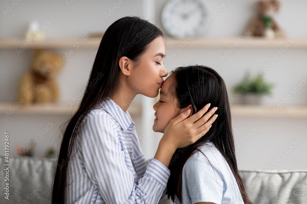 Mother's love. Loving asian mom kissing her daughter in forehead while spending time together at home, side view