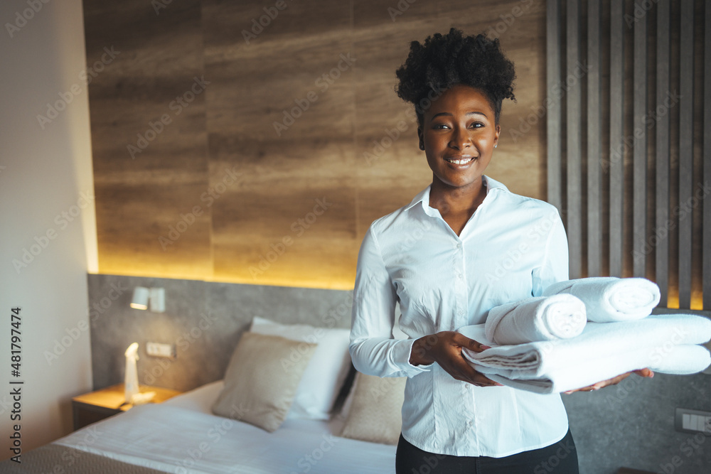 Hotel staff setting up pillow on bed. African housekeeper looking at the camera holding a towel a hotel room. Clean towels during housekeeping in a hotel room Hotel staff setting up pillow on bed. African housekeeper looking at the camera holding a towel a hotel room. Clean towels during housekeeping in a hotel room