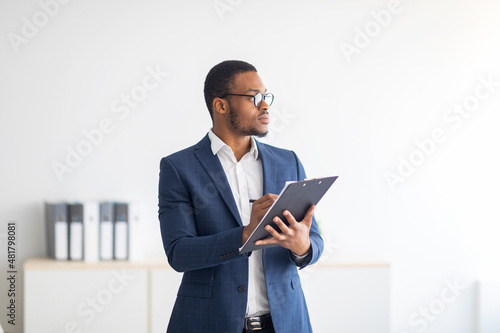 Serious young black psychologist writing in clipboard, looking aside deep in thought at modern office