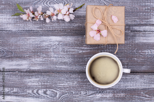 Spring composition with coffee, a gift wrapped in craft paper, spring blossoming almond branches with flowers on a gray shabby wooden table. Flat lay, copy space