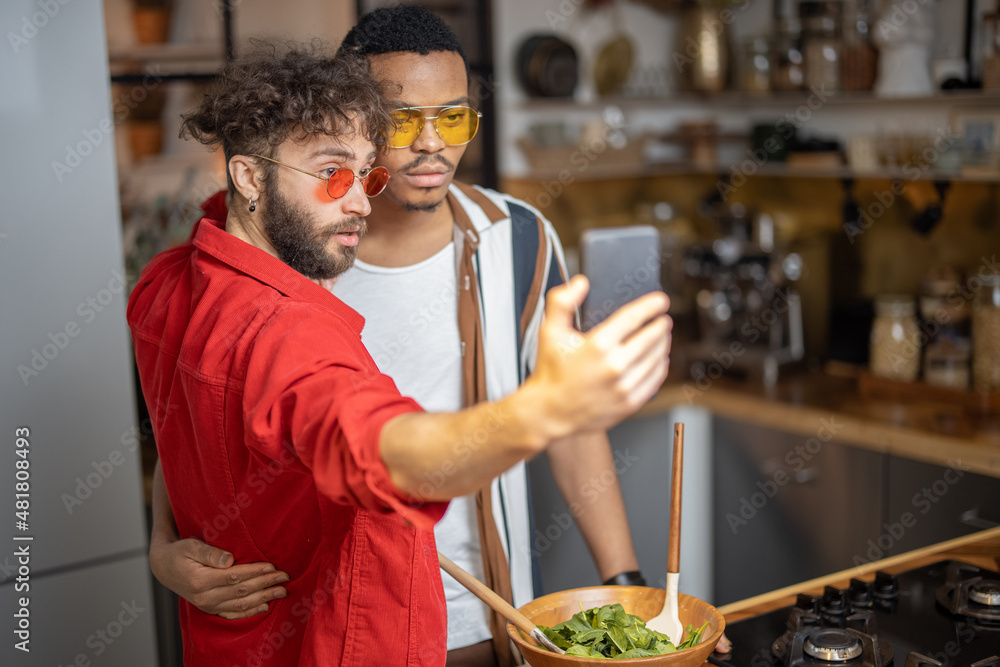 Two brightly dressed stylish guys taking selfie on phone, while having fun together at home. Concept of multiracial diverse gay couple and style. Caucasian and hispanic man having close relationship