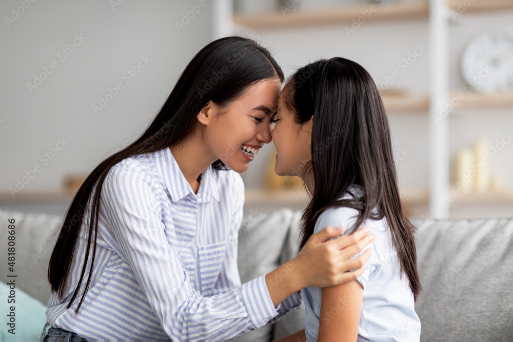 Cute asian mother and daughter cuddling and touching each other with foreheads while sitting on sofa in living room Cute asian mother and daughter cuddling and touching each other with foreheads while sitting on sofa in living room