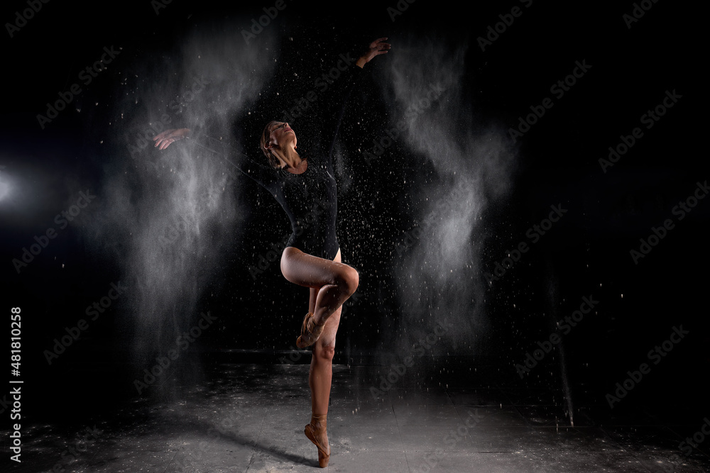 beautiful slender ballet dancer women wearing black bodysuit, sensually poses among flying flour which covers her body, on black studio background. Artistic, commercial, monochrome design.