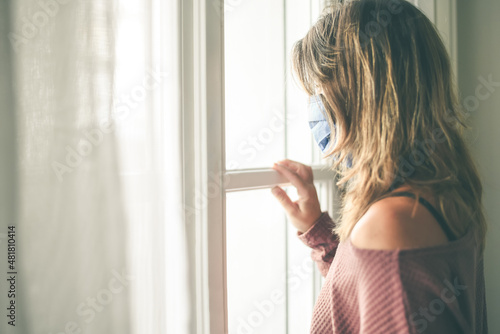 Young woman looking outside the window with surgical mask during coronavirus lockdown Virus covid-19 isolated people at home. Teen waiting to come back to the normal life Girl looks through the glass