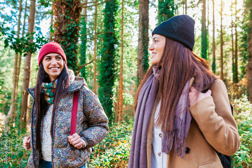 Two cheerful hitchhiker female traveler walking throught winter woodland with backpack. Two smiling beautiful caucasian sisters hiking on the forest looking each other. Tourism and travel concept. photo