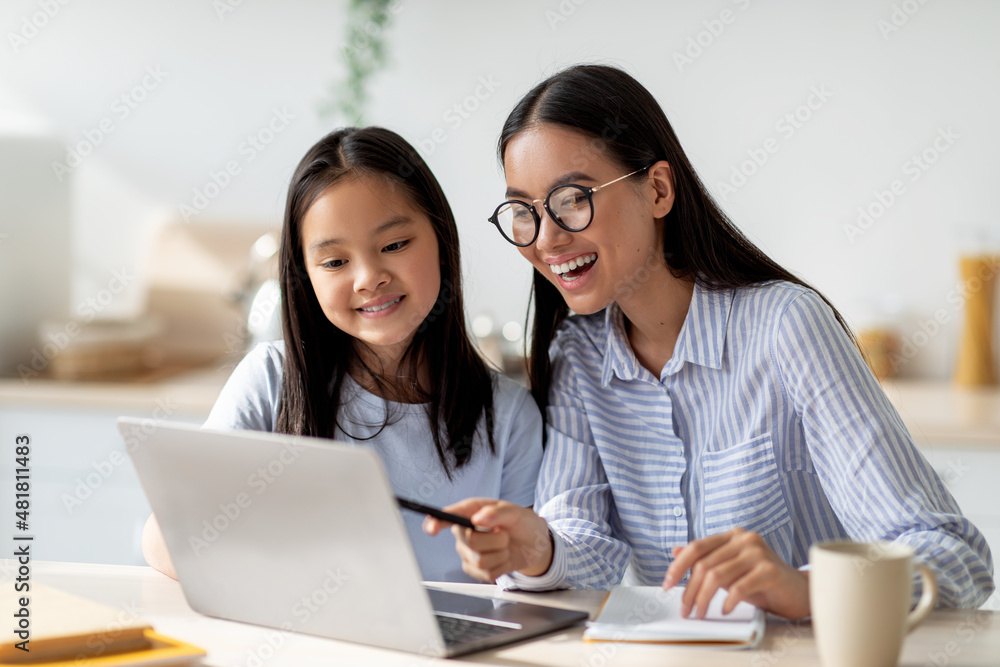 Happy asian mom helping her studying daughter, looking at laptop screen together, sitting in kitchen interior Happy asian mom helping her studying daughter, looking at laptop screen together, sitting in kitchen interior