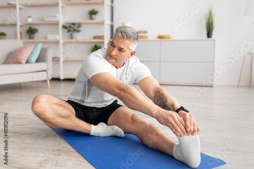 Active lifestyle. Sporty mature man stretching her leg, working out on yoga mat in living room, copy space