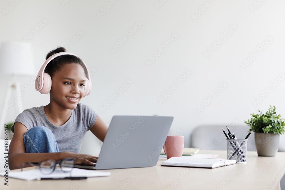 Smiling young black schoolgirl in headphones watch video lesson remotely at laptop at home interior Smiling young black schoolgirl in headphones watch video lesson remotely at laptop at home interior