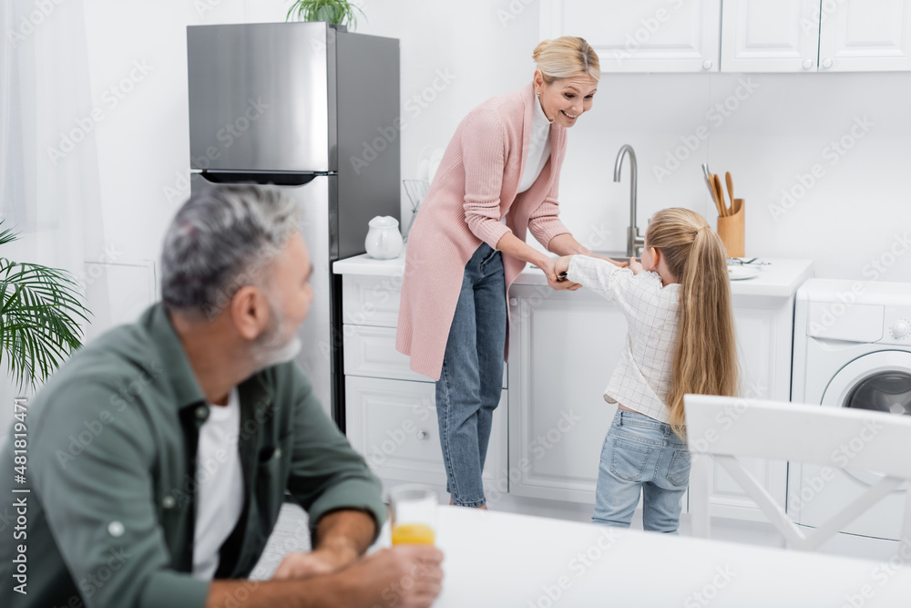 senior man with orange juice looking at granddaughter helping granny in kitchen. senior man with orange juice looking at granddaughter helping granny in kitchen.
