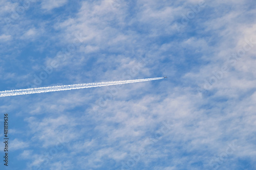 Distant passenger jet plane flying on high altitude through white clouds on blue sky leaving white smoke trace of contrail behind. Air transportation concept