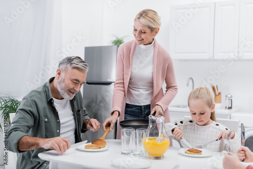 happy woman serving pancakes for husband during breakfast with grandchildren.